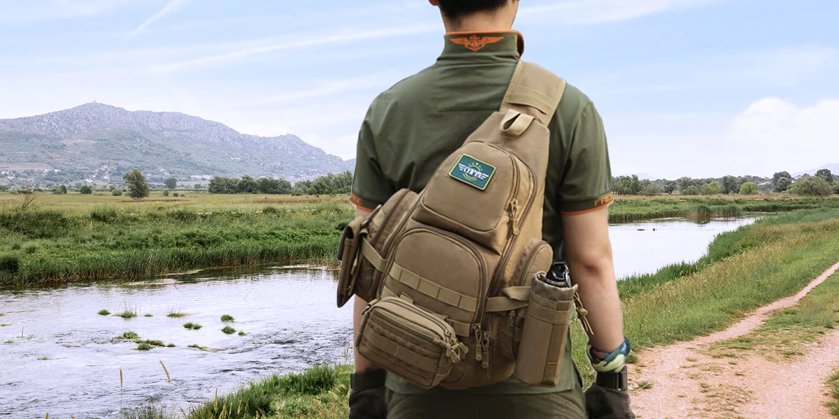 Person wearing a tactical sling backpack in an outdoor setting near a river and mountains.